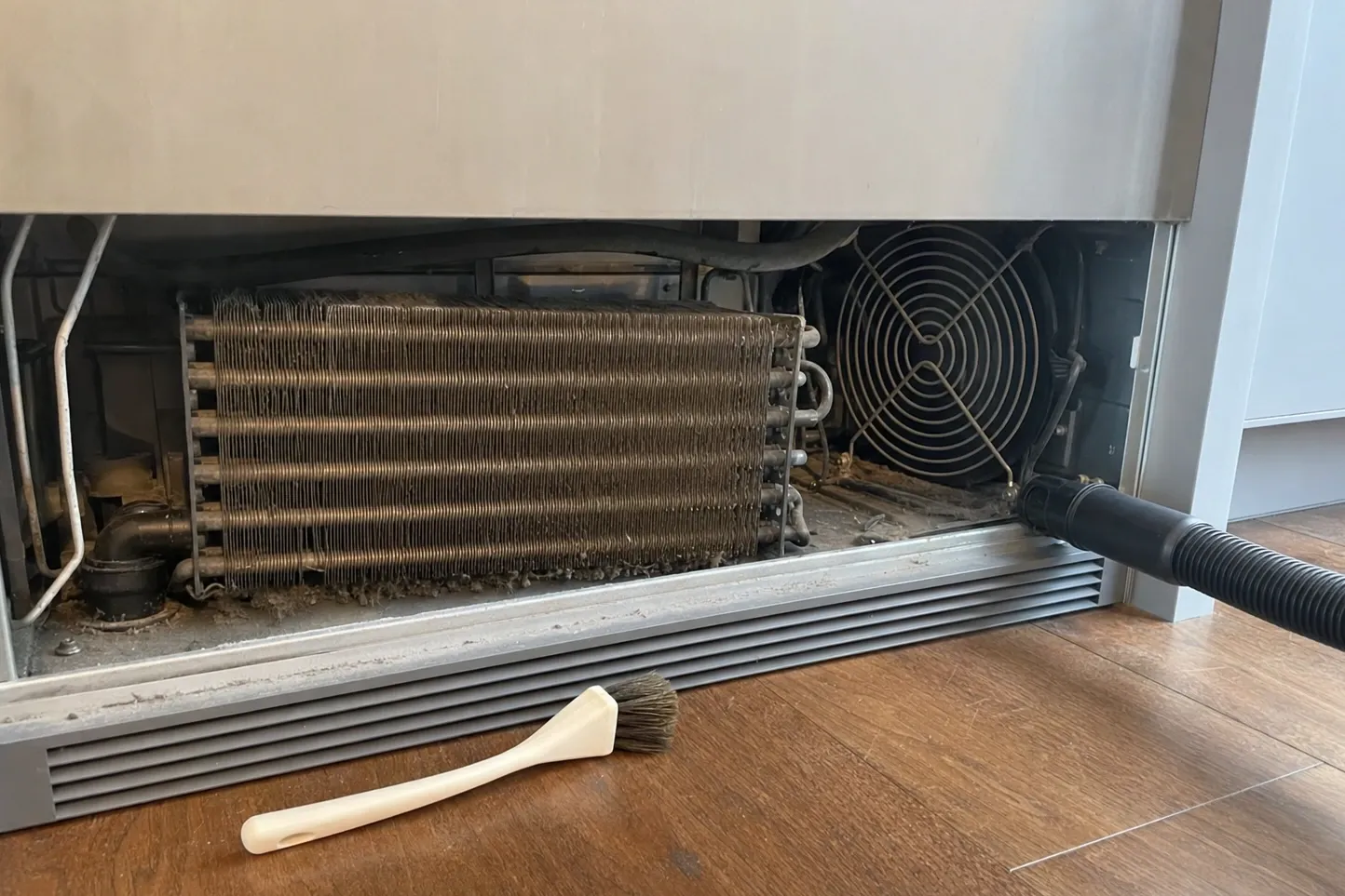 Lower grille removed from a built-in: condenser coil matted grey with dust, condenser fan beside it, vacuum tool and brush ready